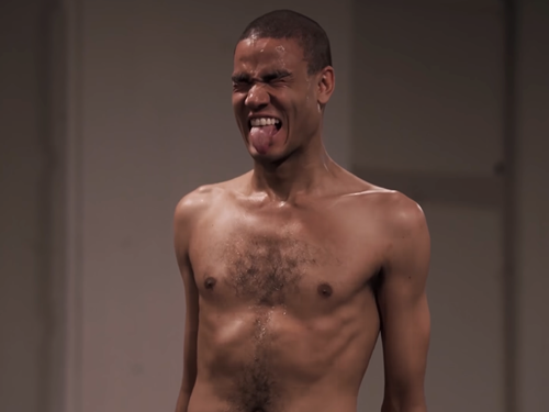 Harald Beharie under fremførelsen av en performance. Han er i halvfigur og bar overkropp. Kroppen er spent og han kniper øynene sammen og rekker tungen ut i en grimase. Fotografi