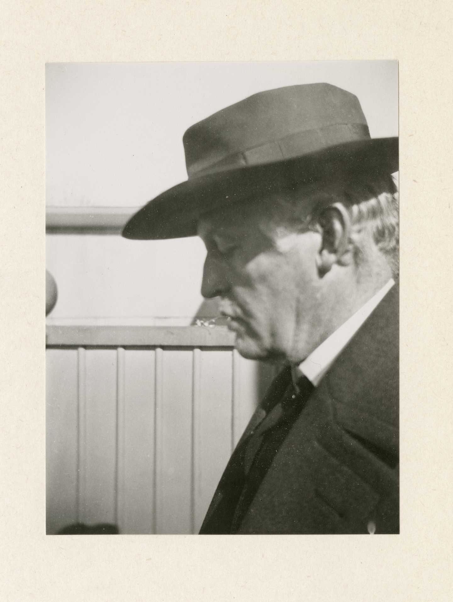 A black and white photograph of Edvard Munch with his face in profile. He wears a suit jacket and a dark hat that casts a shadow over his closed eyes.