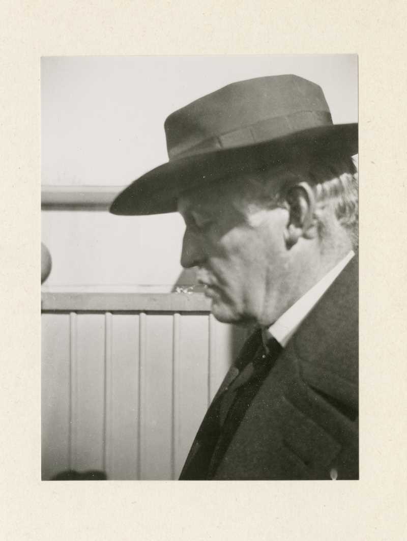 A black and white photograph of Edvard Munch with his face in profile. He wears a suit jacket and a dark hat that casts a shadow over his closed eyes.