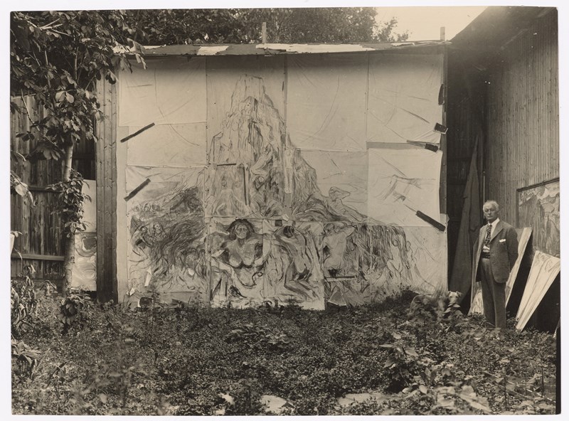 Munch’s open-air studio at Ekely. Edvard Munch is seen on the right side of the image, standing in front of the painting The Human Mountain. Photograph.