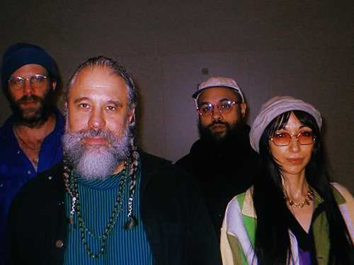 Foto av fire bandmedlemmer. Tre menn med skjegg og én kvinne med langt hår, hatt og solbriller.