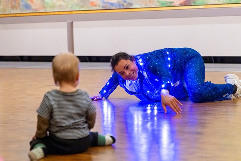I forgrunnen, ute av fokus: en baby som sitter med ryggen til og iakttar en danser, i fokus, litt lengre bak i bildet, som delvis ligger dansende på gulvet og smiler mot barnet. Det blåglitrende kostymet til danseren gir gjenskinn i gulvet.