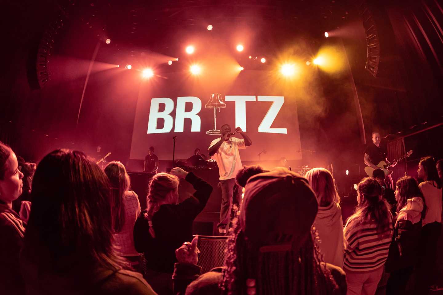 Mørk konsertsal med rødt scenelys, masse publikum og en artist på scenen. Fotografi