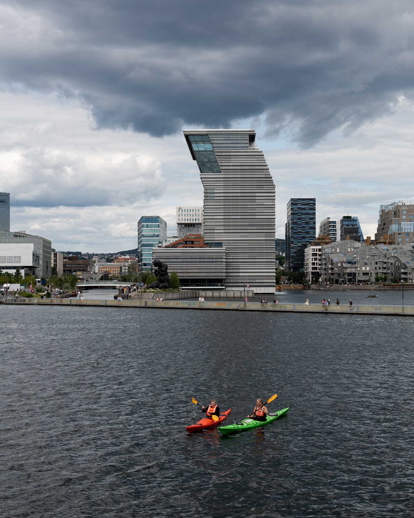 MUNCH sett fra Sørenga. I forkant ser du to mennesker som padler kajakk og bak dem en gangbro som ligger på vannet. Himmelen er blågrå og skyet. Fotografi