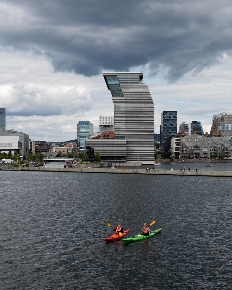 MUNCH sett fra Sørenga. I forkant ser du to mennesker som padler kajakk og bak dem en gangbro som ligger på vannet. Himmelen er blågrå og skyet. Fotografi