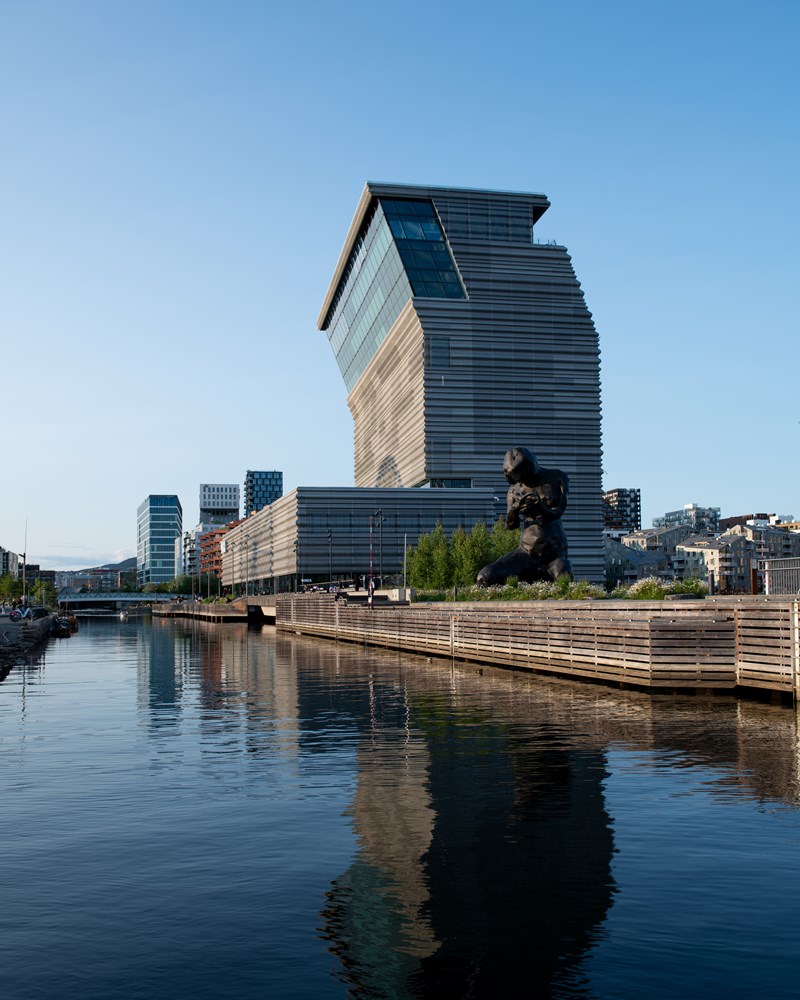 MUNCH-bygget sett fra sørsiden i strålende solskinn. Bygget speiler seg i vannet og du ser skulpturen The Mother i forkant av bildet. Fotografi