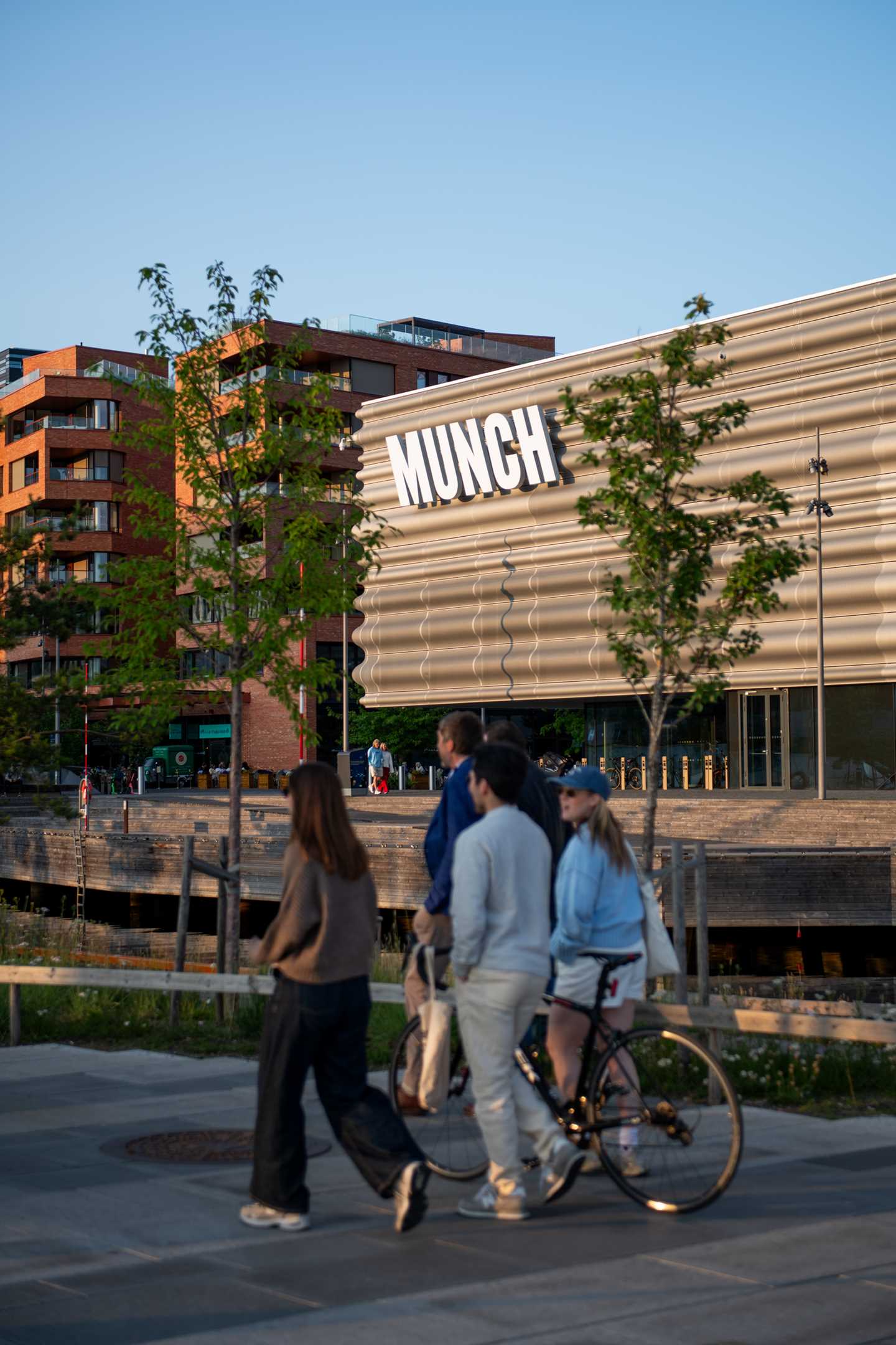 Del av MUNCH-bygget sett fra vest. I forkant går en vennegjeng på fire med en sykkel, i bakgrunnen er bygget med MUNCH-logoen i fokus mellom to grønne trær. Fotografi