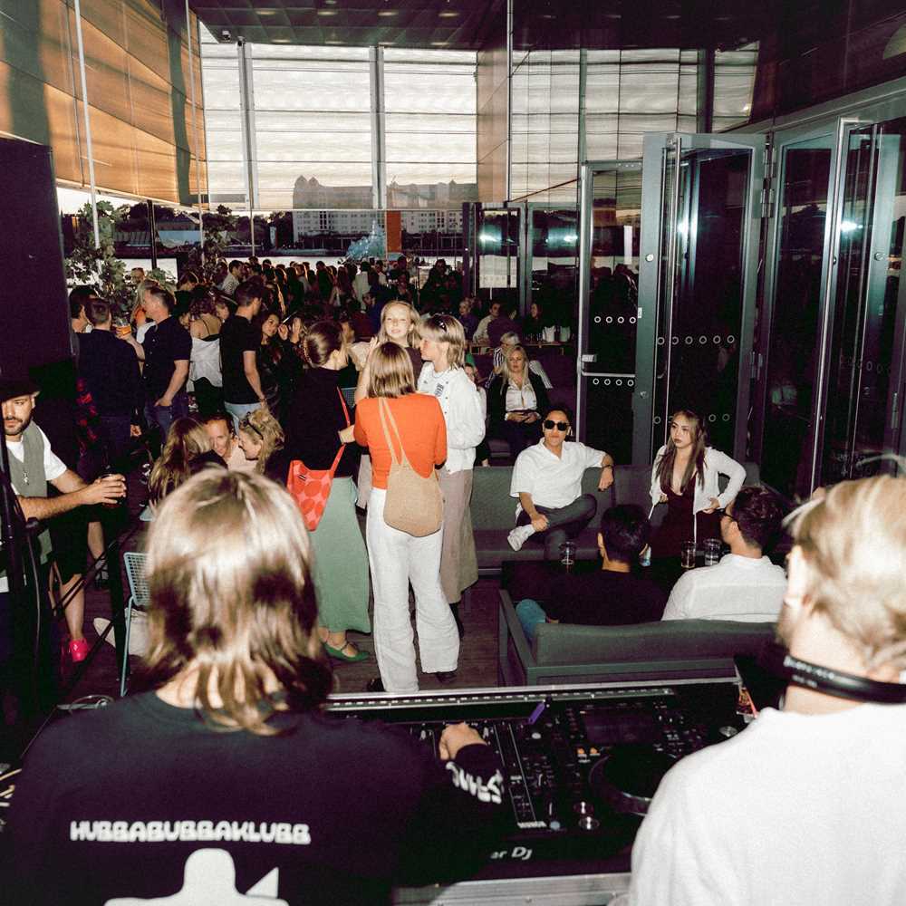 Bakhodene til to DJer skuer ut over menneskemengde på uteservering i kveldssol.