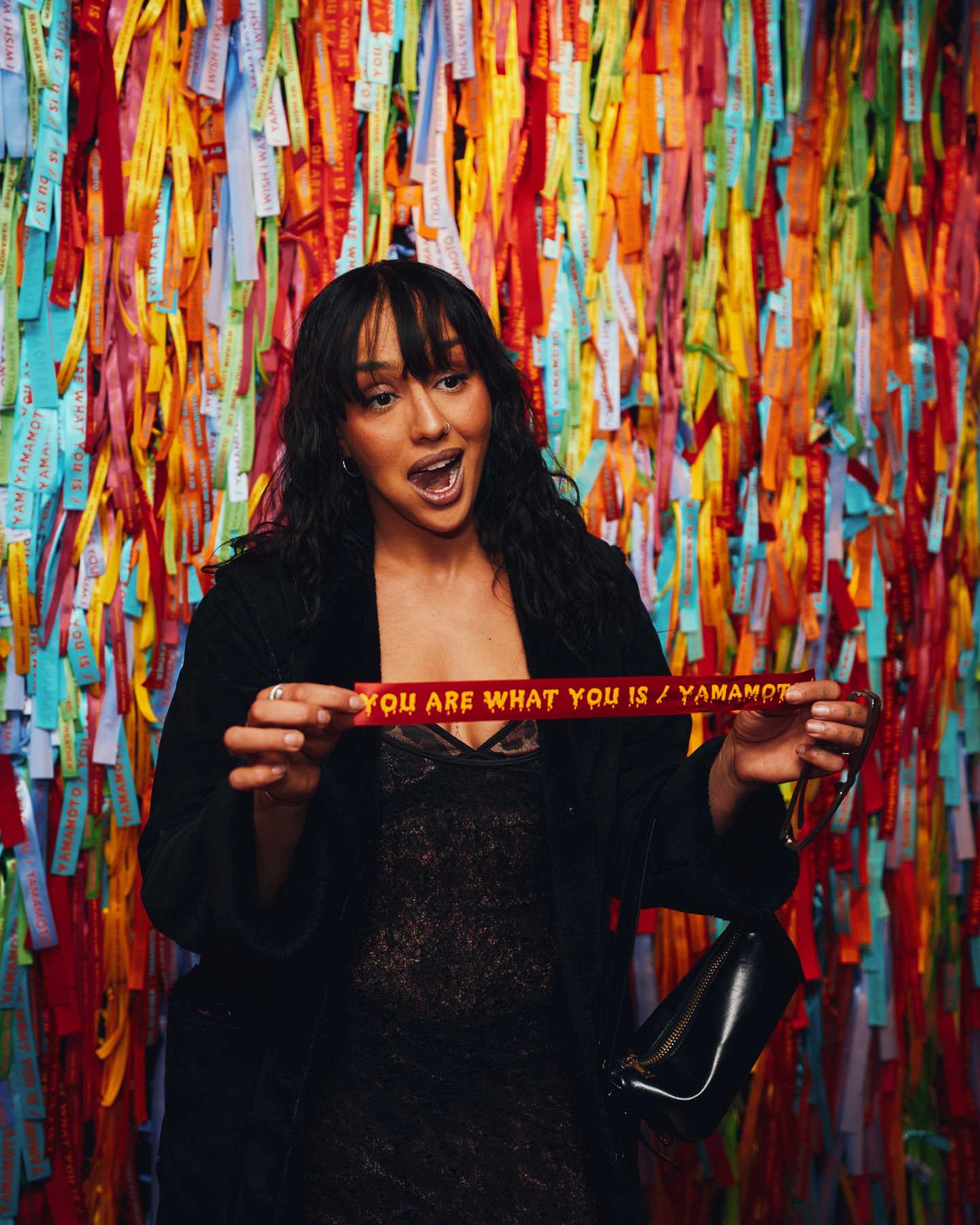 En kvinne står foran en vegg med silkebånd. Hun har tatt et av silkebåndene ned fra veggen og står med det i hånden. Fotogafi