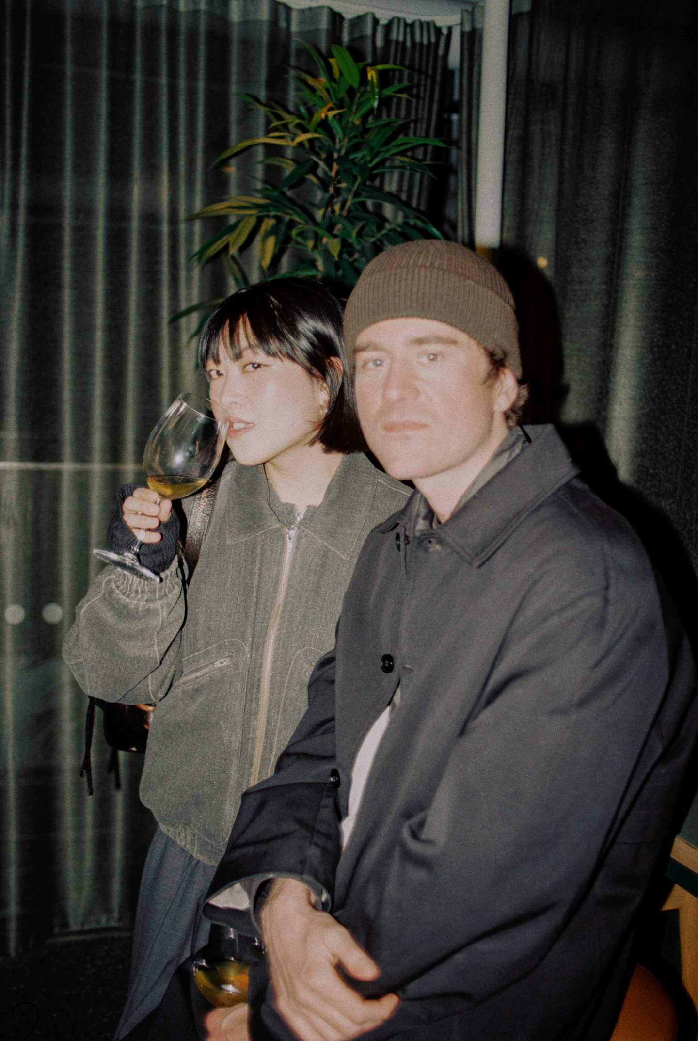 En jente og en gutt sitter i baren på MUNCH med hvert sitt glass hvitvin. Begge ser rett inn i kameraet. Fotografi