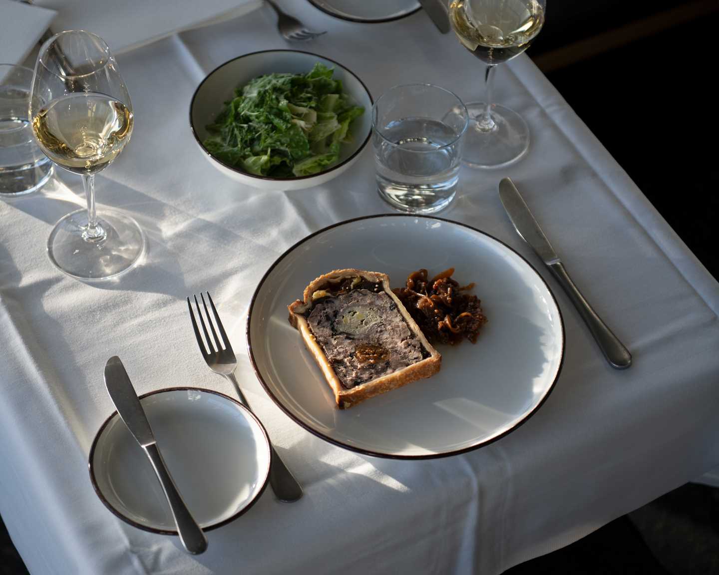 Et bord dekket for to med hvit duk. To glass med hvitvin, en grønn salat står i en bolle midt på bordet og på den ene kuverten er det en tallerken med en paté. Fotografi