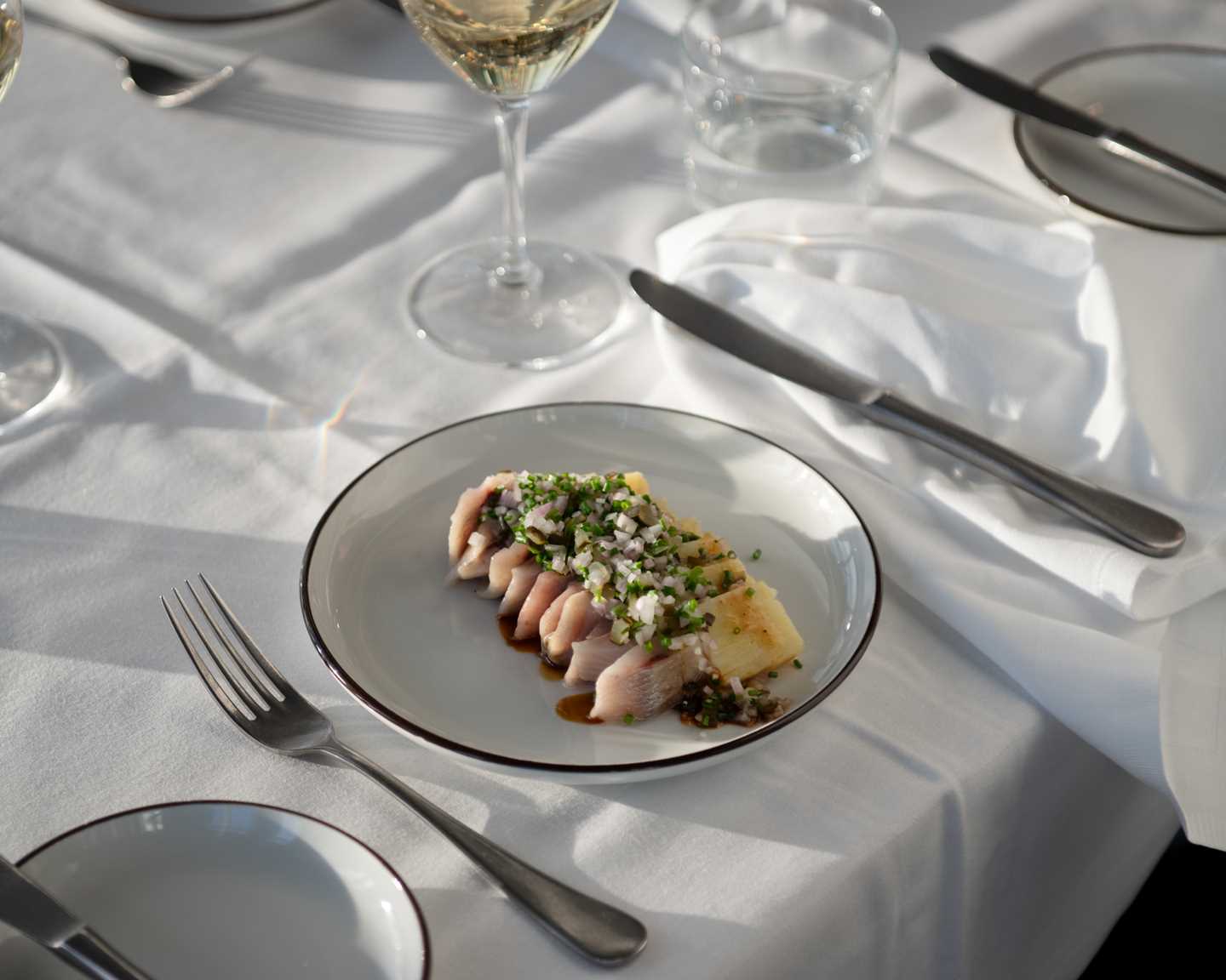 Et bord dekket med hvit duk og stettglass. Nærmest kamera er en tallerken med jomfrusild og poteter, elegant dandert. Ved siden av står et vinglass med hvitvin. Fotografi