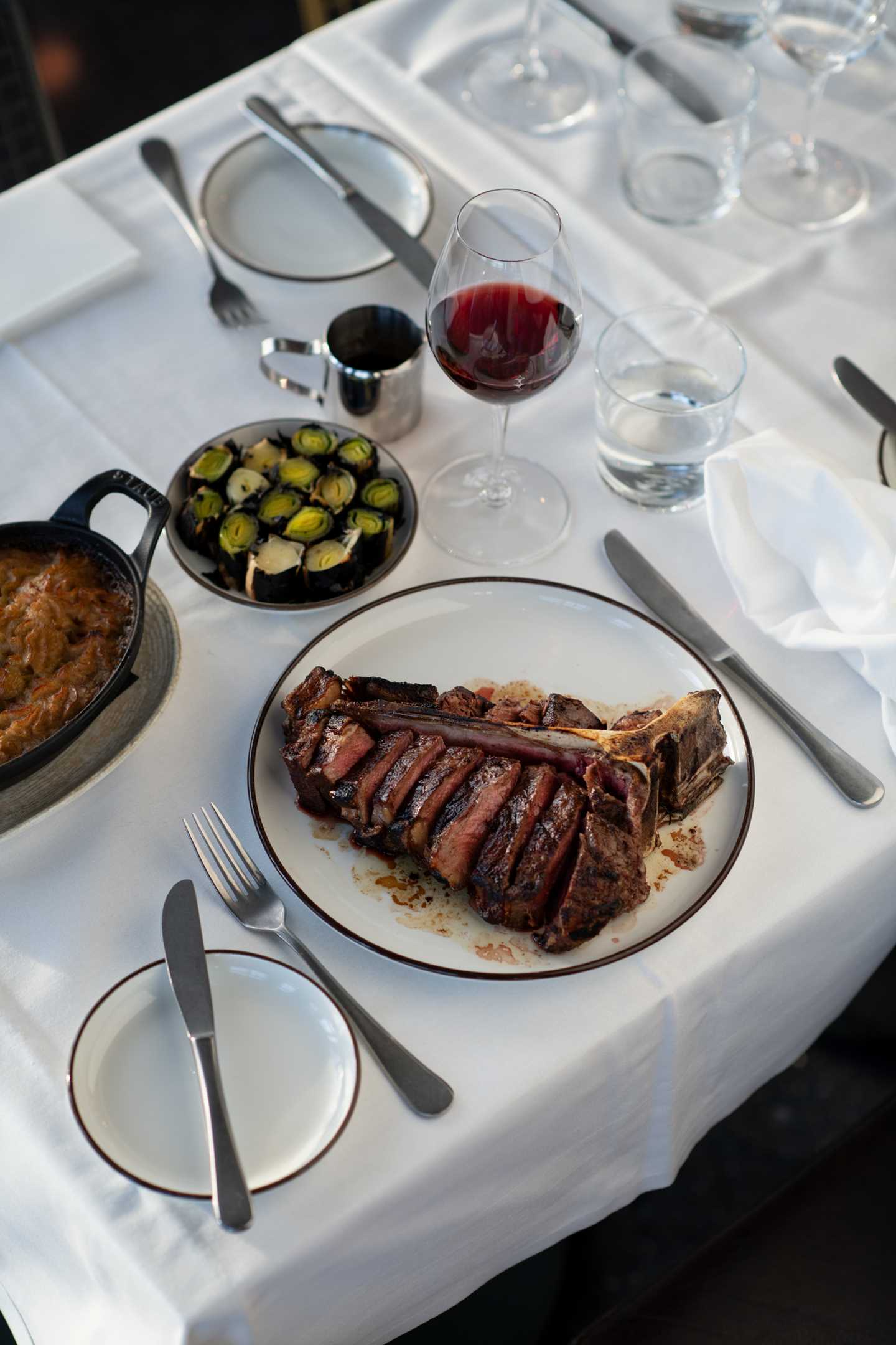 Et bord dekket med hvit duk, bestikk og stettglass. I fokus er en tallerken med en kraftig t-bone-steak. Ved siden av står et vinglass med rødvin, en tallerken med bakte purrebiter og en ildfast form poteter. Fotografi