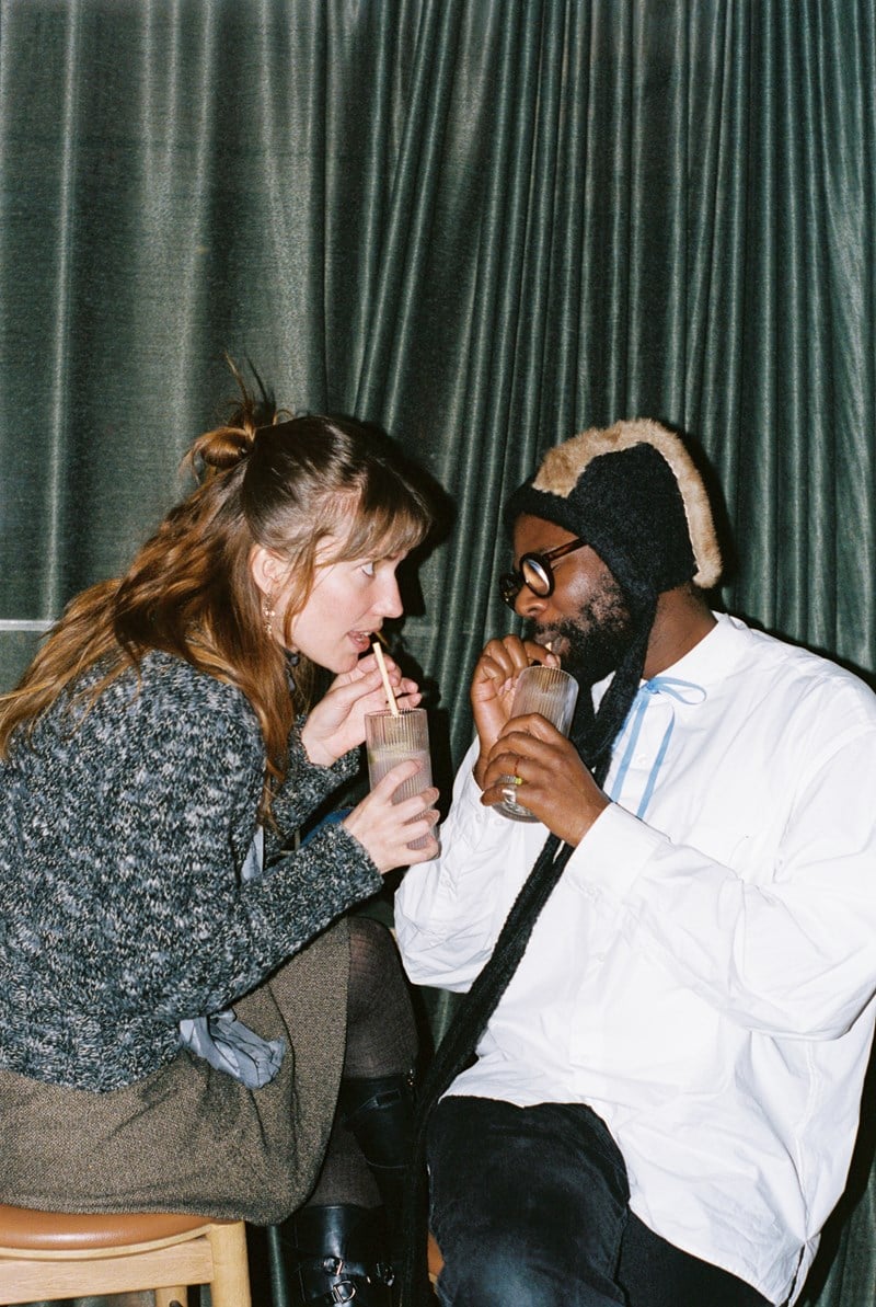 En kvinne og mann sitter tett, vendt mot hverandre og drikker av hver sin drink med sugerør mens kvinnen stirrer intenst på mannen, som har lukkede øyne. Fotografi