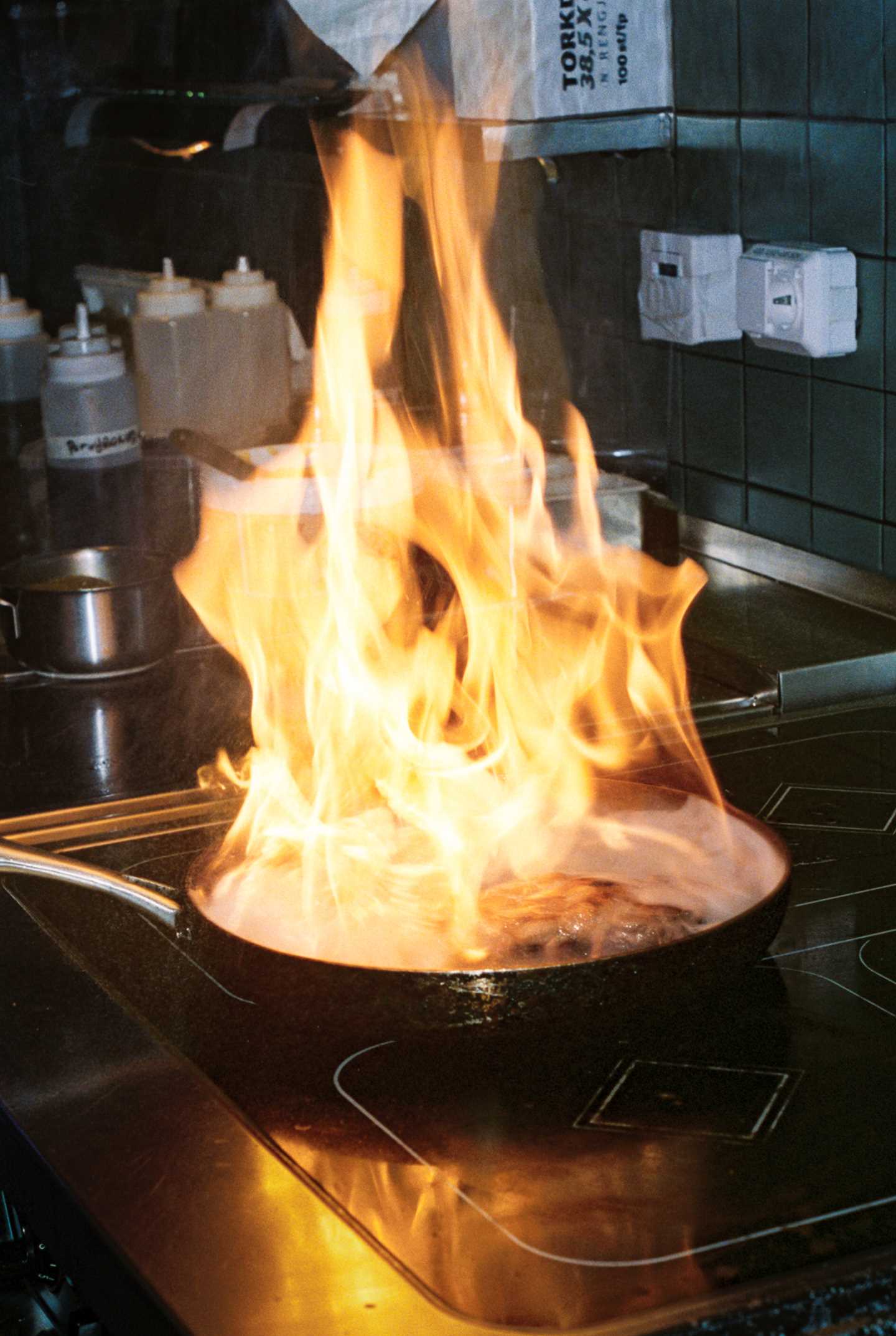 Store flammer stiger opp fra en stekepanne der noe blir flambert på et restaurantkjøkken. Fotografi