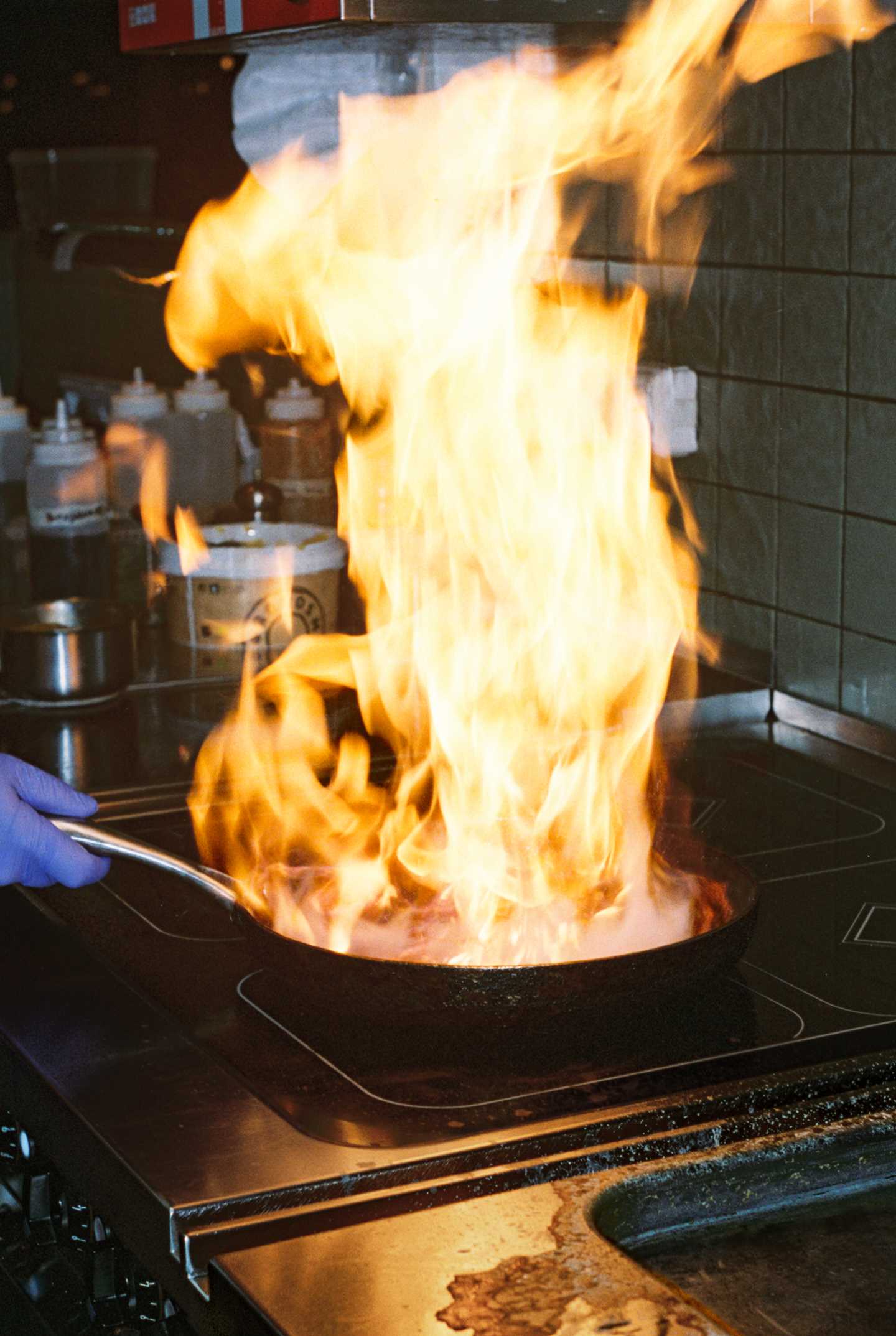 Store flammer stiger opp fra en stekepanne der noe blir flambert på et restaurantkjøkken. Foruten flammene er det en hånd med blå plasthanske som holder håndtaket på stekepannen og i bakgrunnen kan vi se flasker med dressinger, oljer og sauser. Fotografi
