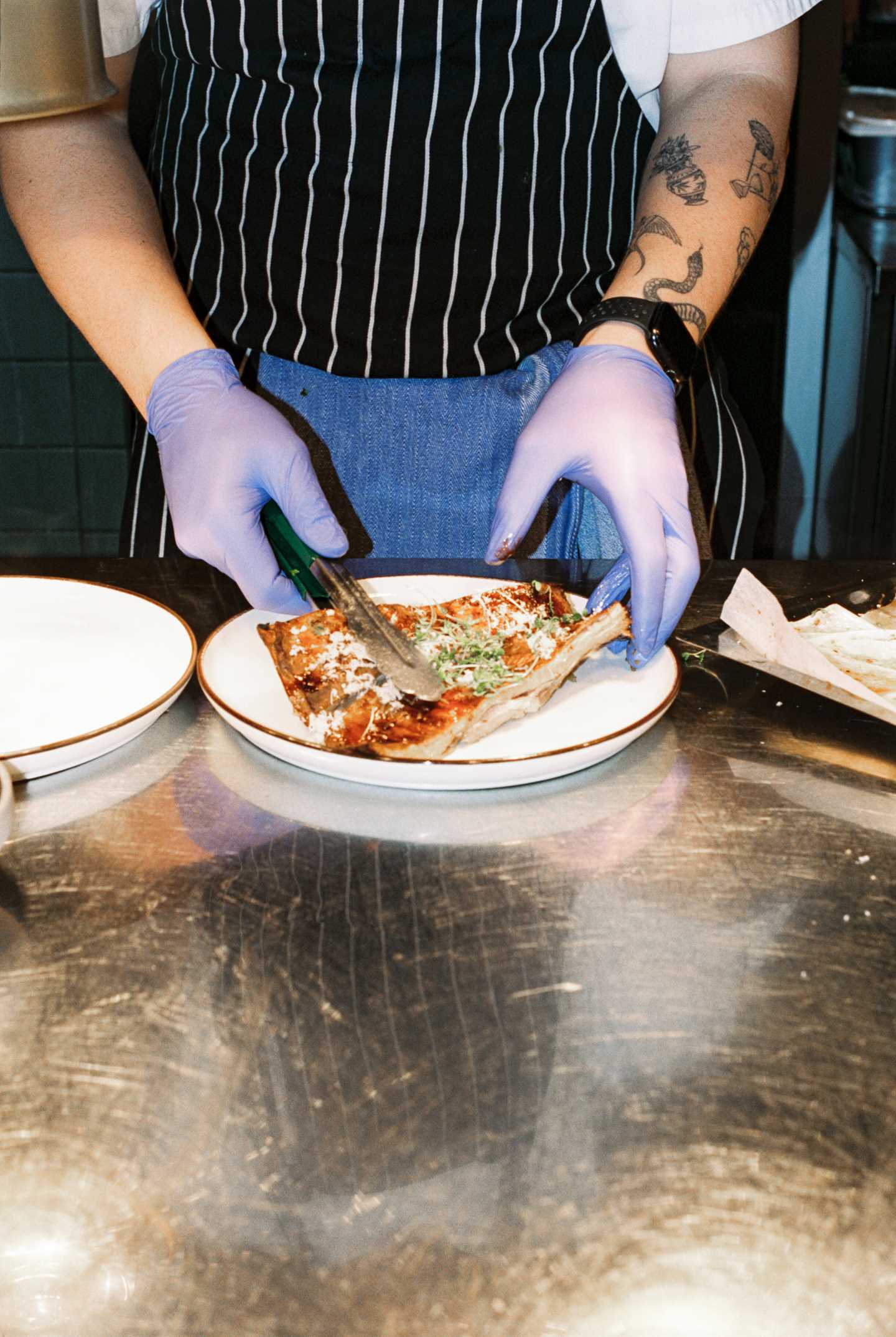 Vi ser armene til en kokk med blå plasthansker på hendene som plasserer et pizzastykke på en tallerken. Fotografi