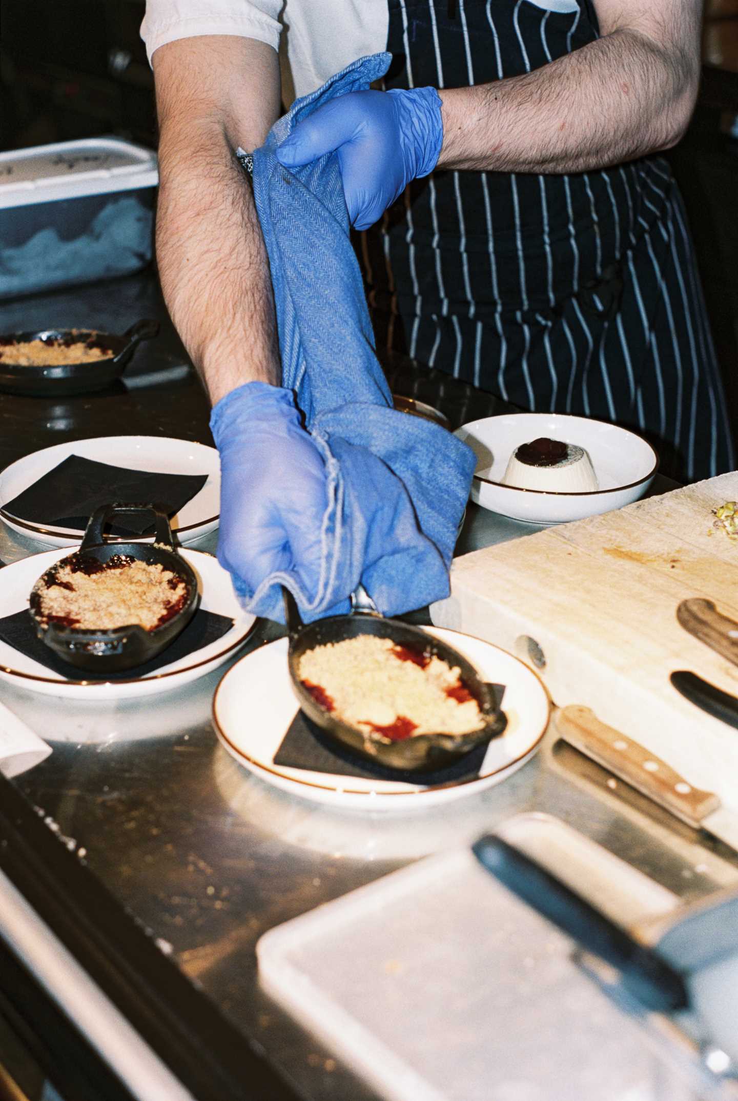 En kokk med blå plasthansker på hendene og stripete forkle løfter en ildfast form fra ovnen til en tallerken. Han holder formen med et kopphåndkle for å ikke brenne seg. Fotografi