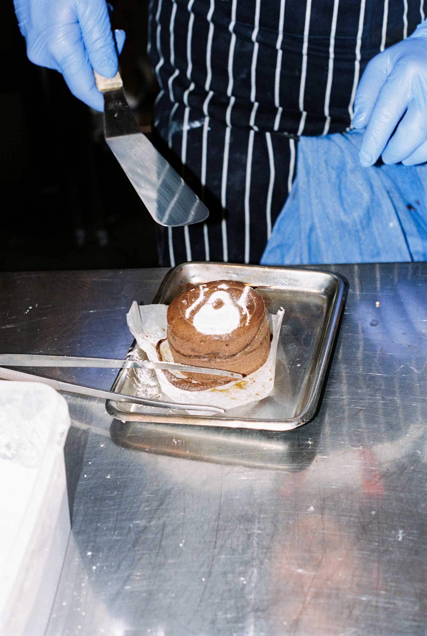 En sjokoladefondant dekorert med melis som er strødd på toppen i form av Skrik-motivet. I bakgrunnen ser vi hendene til en kokk med stekespade. Fotografi