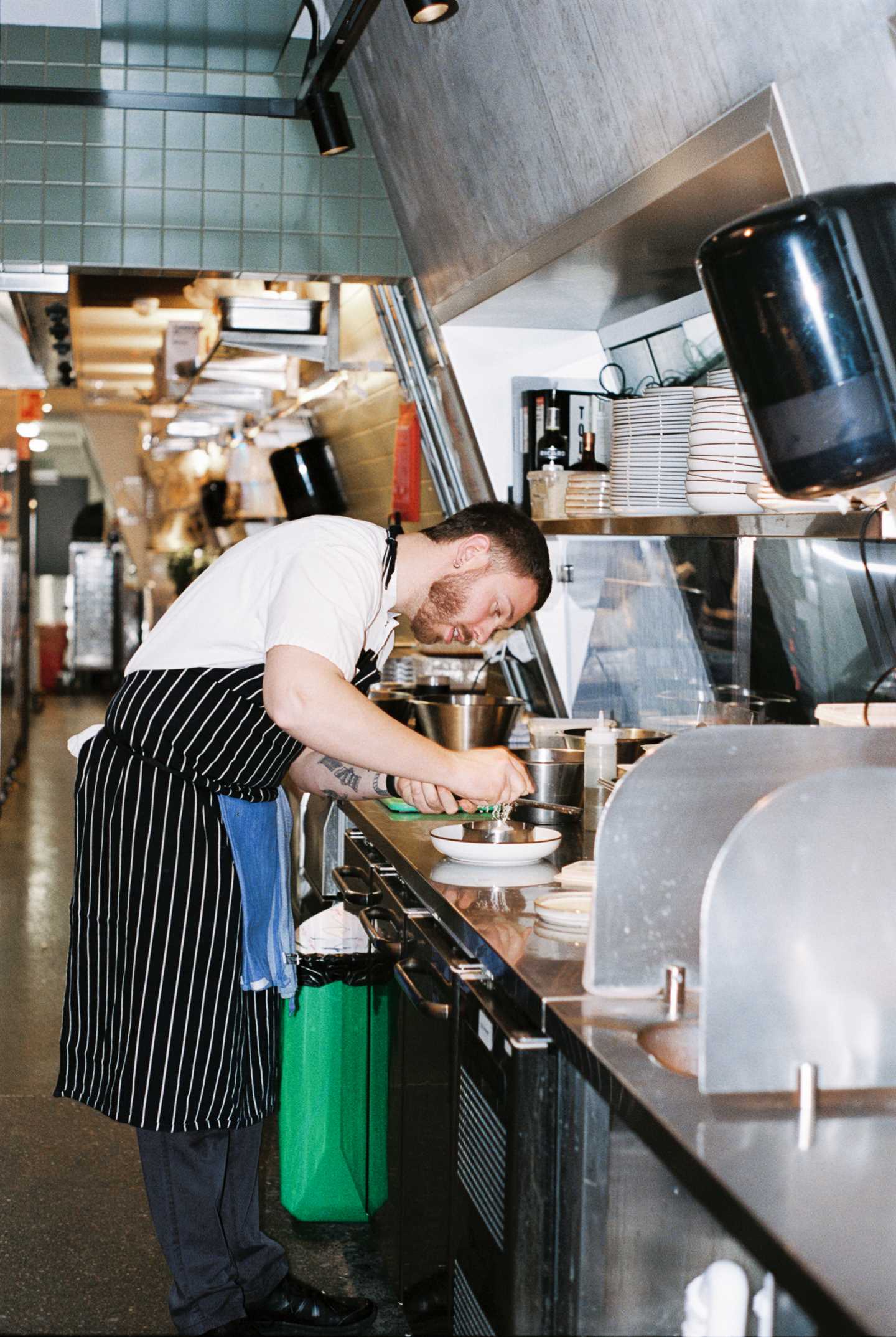 En kokk står bøyd over en kjøkkenbenk på et restaurantkjøkken. Han river noe hvitt på et rivjern over en rett. Fotografi