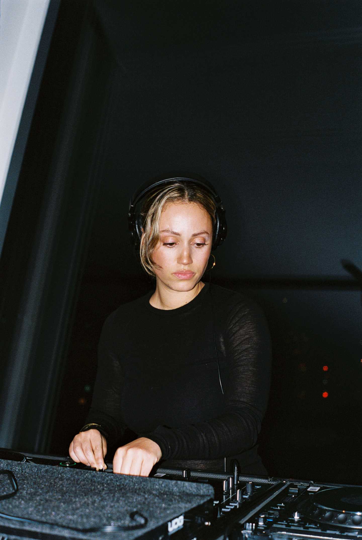 En DJ kledd i svart med blondt hår og headset står bak en miksepult foran et stort vindu. Utenfor vinduet er en nattsvart himmel. Fotografi