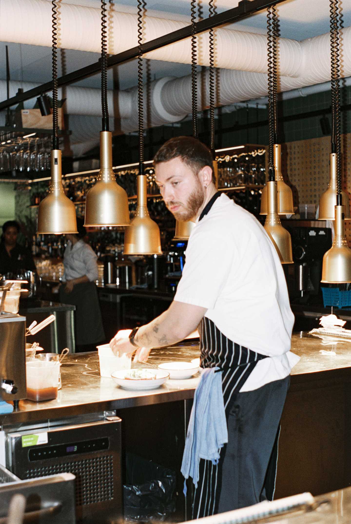 Et restaurantkjøkken med en kjøkkenbenk med varmelamper hengende ned fra taket over. Ved kjøkkenbenken står en kokk, kledd i hvit t-skjorte og stripete forkle og danderer en rett før servering. Fotografi