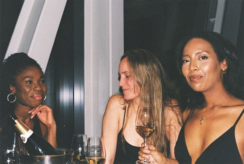 Tre kvinner sitter og står rundt et bord med flasker og glass på. Kvinnen til høyre i bildet ser rett i kamera, mens kvinnen i midten ser ned mot gulvet og kvinnen til venstre i bildet ser på kvinnen som ser inn i kameraet. Fotografi