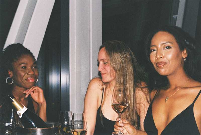 Three women sit and stand around a table with bottles and glasses. The woman on the right looks directly at the camera, the woman in the middle looks down at the floor, and the woman on the left looks at the woman facing the camera. Photograph.