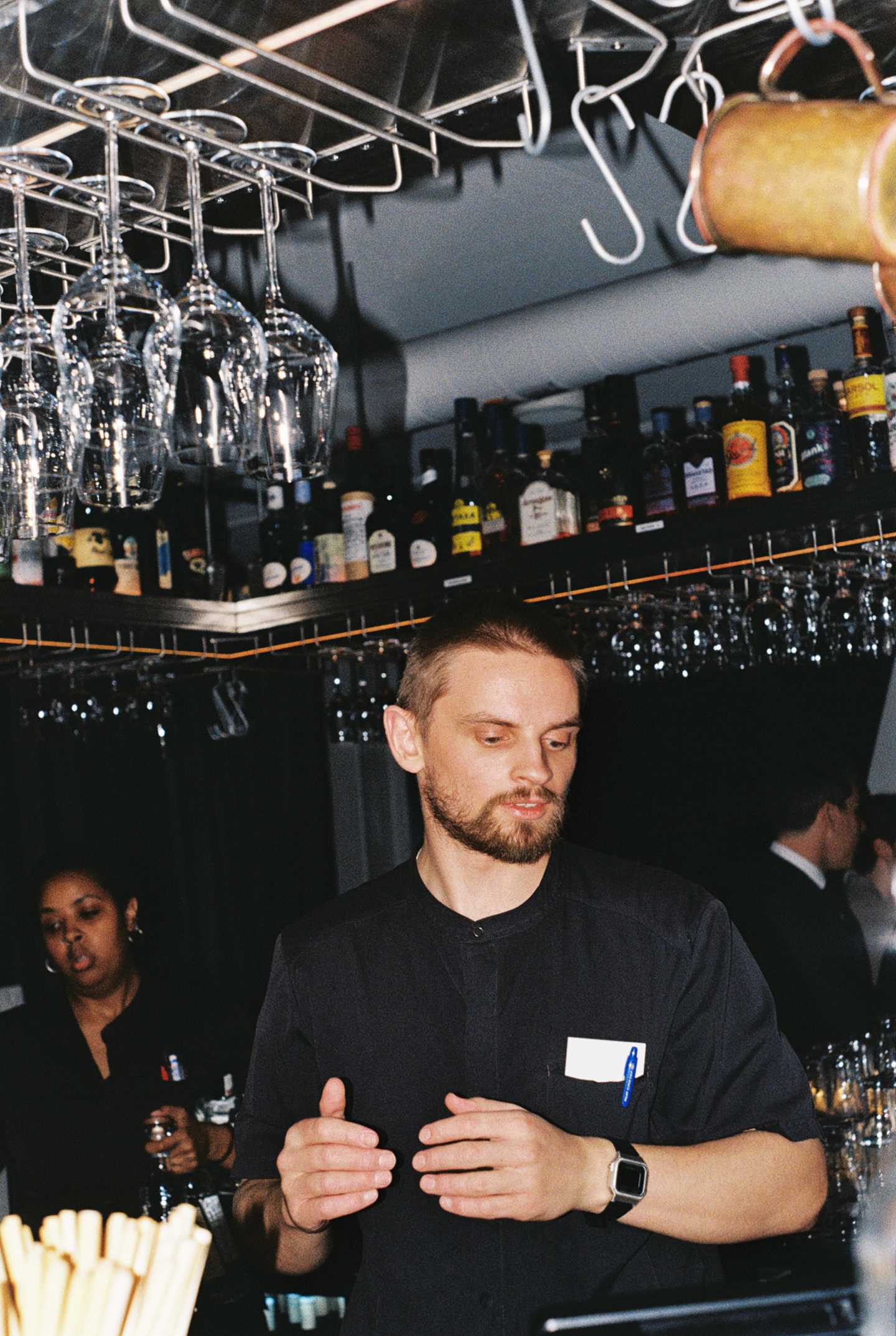 En bartender står i midten av en bardisk som går rundt ham i en firkant. På en hylle oppunder taket ser vi ulike spritflasker og under hyllen henger stettglass ned fra stativer. Fotografi