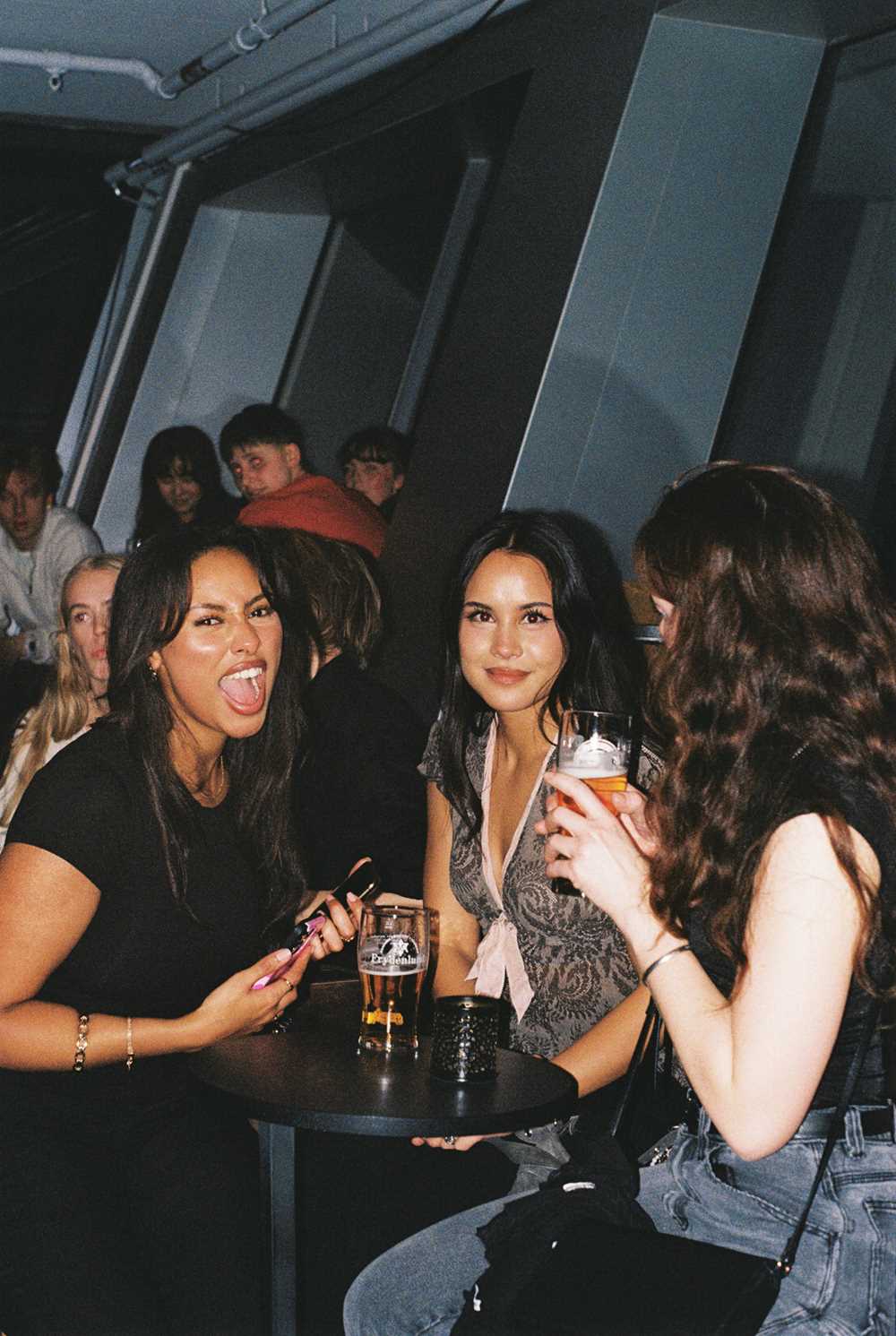 Tre jenter står og sitter rundt et lite rundt bord mens de drikker hver sin øl. En av jentene ser bort fra kameraet, de to andre ser rett på kameraet og jenta lengst til venstre i bildet åpner munnen som i et brøl. I bakgrunnen ser vi mange andre bargjester. Fotografi