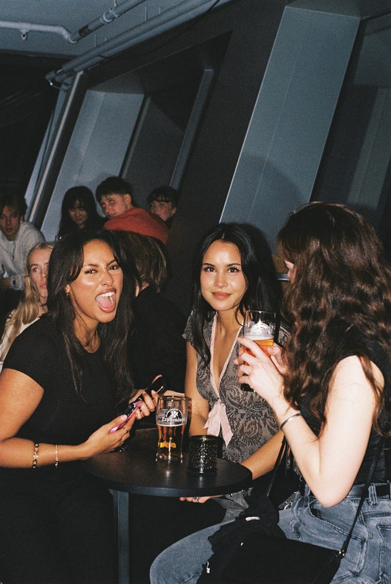 Tre jenter står og sitter rundt et lite rundt bord mens de drikker hver sin øl. En av jentene ser bort fra kameraet, de to andre ser rett på kameraet og jenta lengst til venstre i bildet åpner munnen som i et brøl. I bakgrunnen ser vi mange andre bargjester. Fotografi