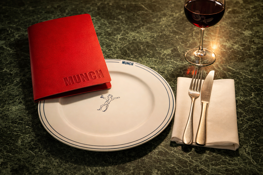 På et grønt marmorbord står en hvit tallerken med en tegning på midten av tallerkenen. Oppå tallerkenen ligger en rød meny. Ved siden av tallerken ligger en hvit serviett med kniv og gaffel på. Ovenfor servietten står et vinglass med rødvin. Fotografi