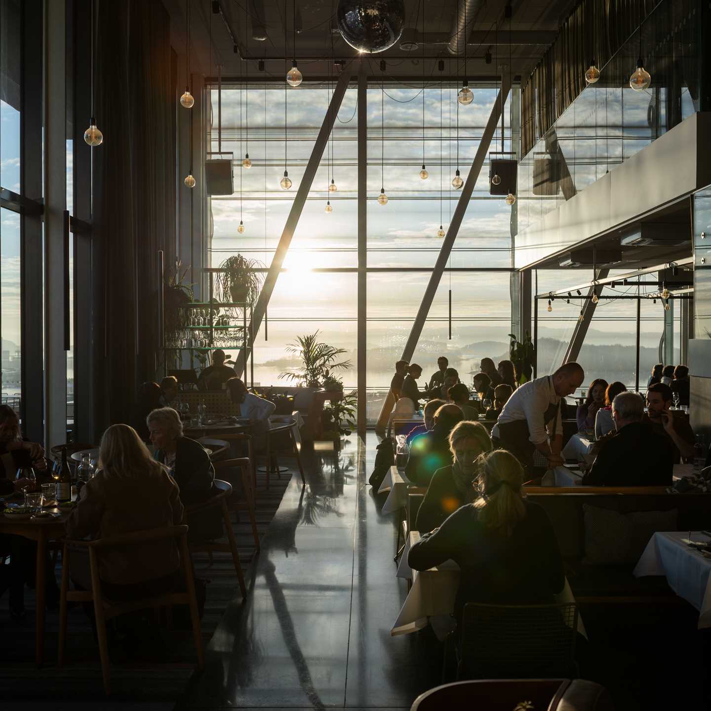 MUNCH restaurant badet i solskinn som kommer inn gjennom veggen av vinduer i enden av lokalet. Vi ser en full restaurant med mennesker ved alle bord og servitører som går rundt mellom bordene. Fotografi