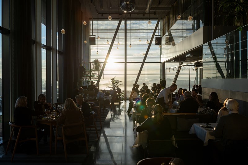 MUNCH restaurant badet i solskinn som kommer inn gjennom veggen av vinduer i enden av lokalet. Vi ser en full restaurant med mennesker ved alle bord og servitører som går rundt mellom bordene. Fotografi