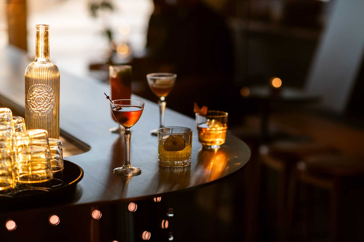 Bardisk med ulike drinker i ulike glass. I kanten av bildet står et brett med tomme glass og en glassflaske fylt med vann. Fotografi