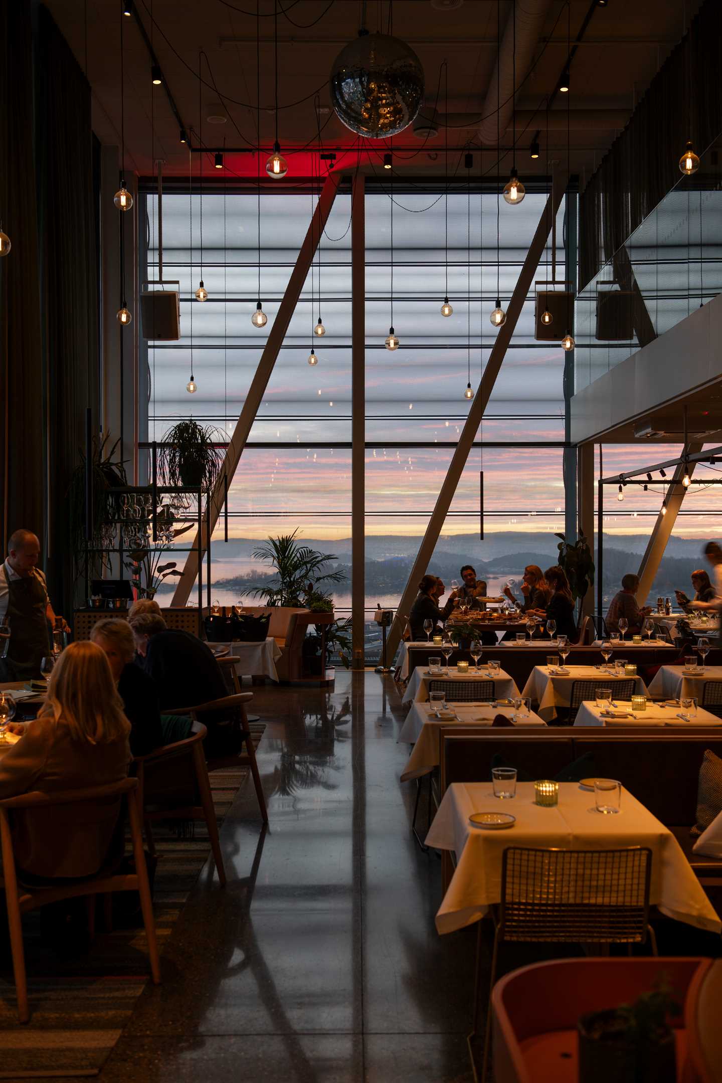 En halvfull restaurant med små og store bord med hvite duker og flere grupper ved ulike bord. I enden av lokalet er det vinduer fra gulv til tak og utenfor ser man solnedgangen og Oslofjorden. Fotografi