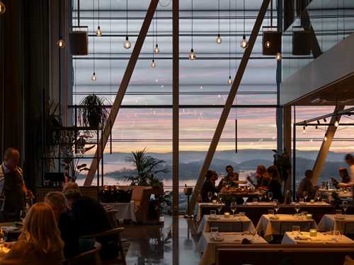 En halvfull restaurant med små og store bord med hvite duker og flere grupper ved ulike bord. I enden av lokalet er det vinduer fra gulv til tak og utenfor ser man solnedgangen og Oslofjorden. Fotografi
