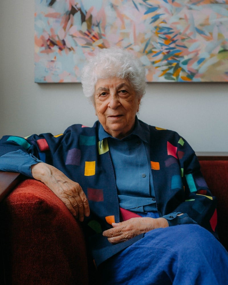 Kunstneren sitter i en sofa med armen over armlenet. Hun ser rett inn i kameraet og bak henne på veggen ser vi et av hennes kunstverk. Fotografi