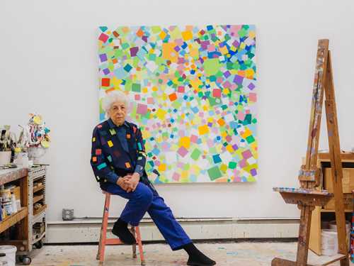 Kunstneren Samia Halaby sitter på en rosa krakk i sitt atelier foran ett av sine egne verk. Veggen er hvit og verket består av mange fargerike firkanter. Til høyre i bildet står et staffeli med malingsflekker og til venstre i bilde ser vi en hylleseksjon som er full av maling og malersaker. Fotografi