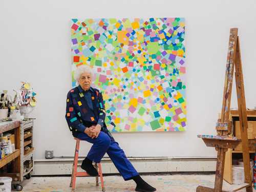 Kunstneren Samia Halaby sitter på en rosa krakk i sitt atelier foran ett av sine egne verk. Veggen er hvit og verket består av mange fargerike firkanter. Til høyre i bildet står et staffeli med malingsflekker og til venstre i bilde ser vi en hylleseksjon som er full av maling og malersaker. Fotografi