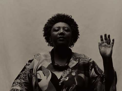 Portrett av en sittende Rosana Paulino. Hun holder venstre hånd løftet som om hun avlegger en ed. Bildet er i svart/hvitt. Fotografi.