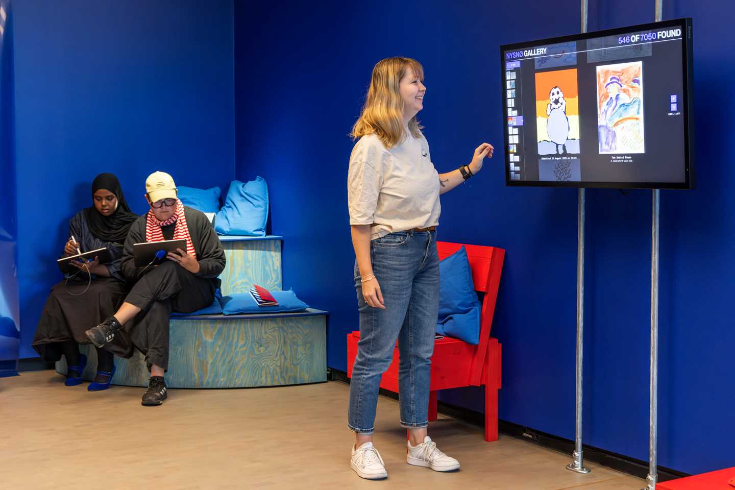 En jente blar gjennom tegninger som er sendt inn digitalt på en skjerm. I bakgrunnen sitter to personer og tegner på nettbrett. Fotografi
