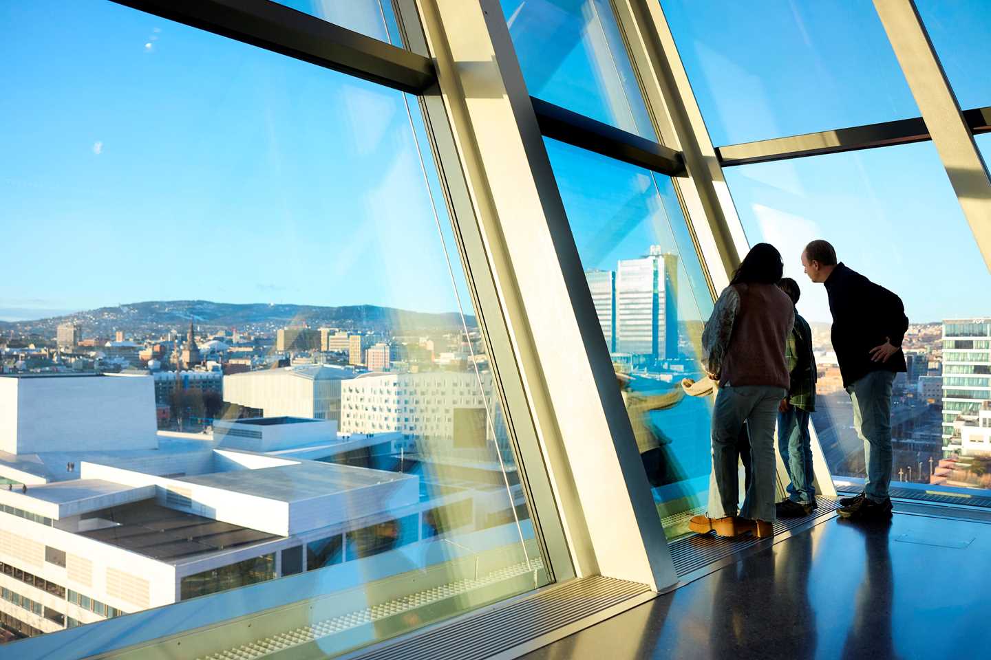 En familie står ved vinduene i 10. etasje på MUNCH og ser utover mot Oslo. Utenfor er det blå himmel og vi ser operahuset. Fotografi