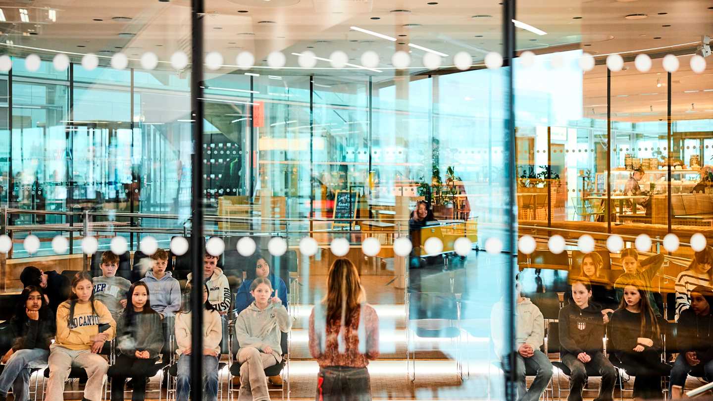 Amfiet på MUNCH sett utenfra. På den andre siden av glasset ser vi en skoleklasse som lytter til en formidler som står midt på gulvet foran elevene. Fotografi