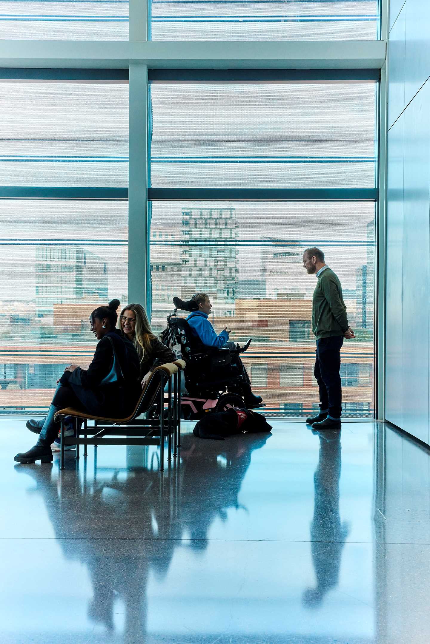 To kvinner sitter og ler på en benk utenfor et utstillingsrom på MUNCH. Bak dem er en kvinne i rullestol i samtale med en mann. På gulvet ved siden av rullestolen ligger en tjenestehund. Fotografi