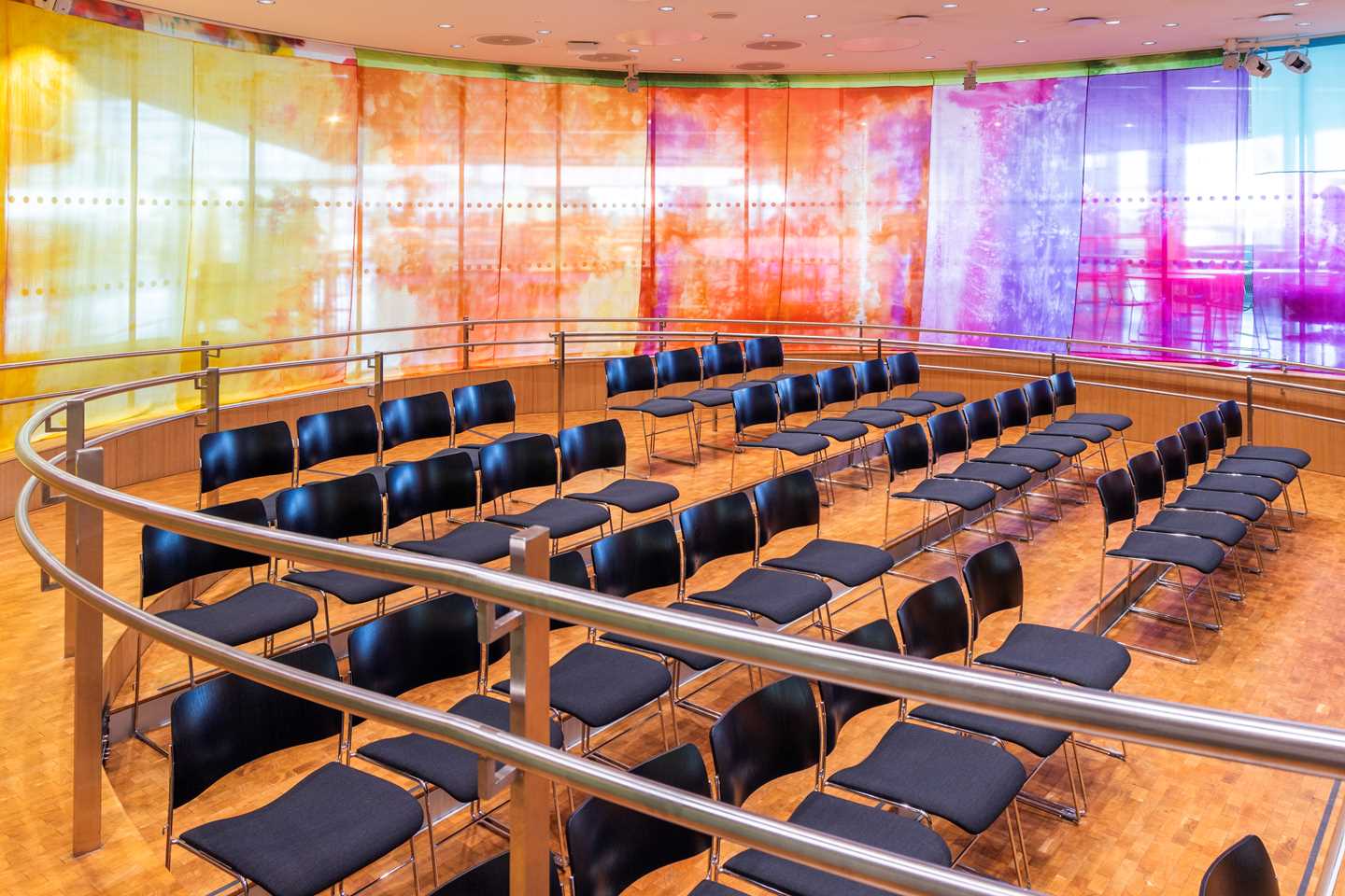 Amifet på MUNCH sett fra inngangen. Et rundt rom med vinduer hele veien rundt. Foran vinduene henger det et tynt silkestoff i alle regnbuens farger. I midten av rommet står det flere rader med svarte stoler. Fotografi