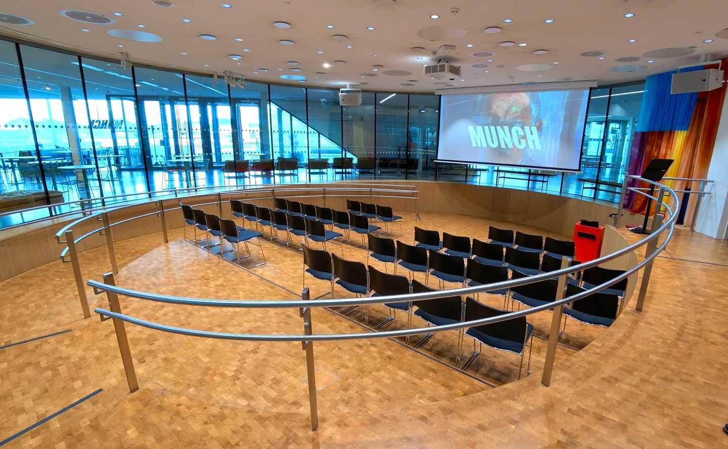 Amfi-salen på MUNCH. Et rundt rom med vinduer rundt hele rommet. I midten av rommet er det fire stolrader som er vendt mot et lerret som henger ned fra taket. Fotografi
