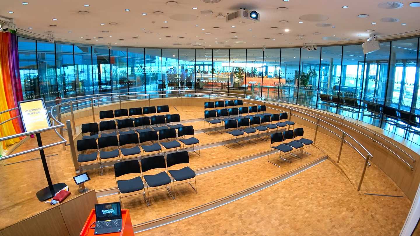 Amfi-salen på MUNCH. Et rundt rom med vinduer rundt hele rommet. I midten av rommet er det fire stolrader som er vendt mot et lerret som henger ned fra taket. Fotografi