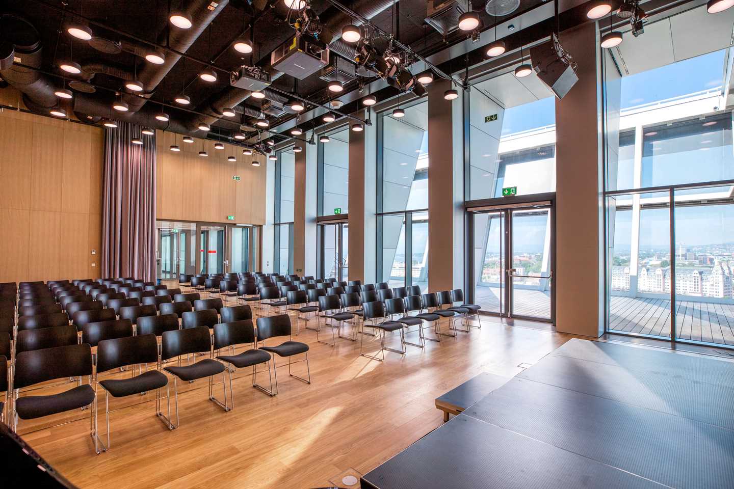 Toppsalen på MUNCH sett fra scenen. Inne i rommet er det rader med svarte stoler og den ene veggen består av vinduer fra gulv til tak og utenfor er det en takterrasse. Fotografi