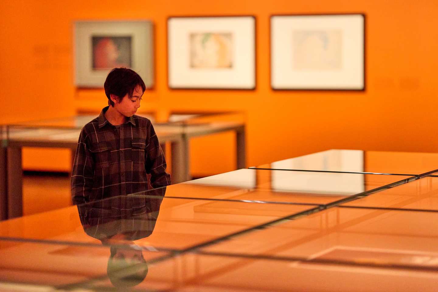 En ung gutt står ved siden av glassmontré og ser på trykk av Edvard Munch i utstillingen Inntil. Fotografi